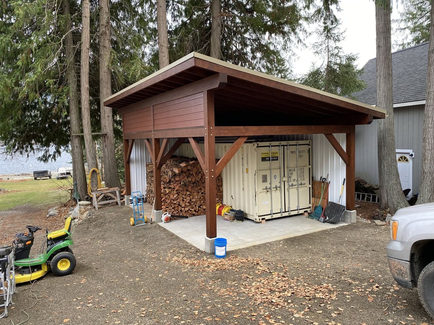Timber post-and-beam shelter with a sloped roof, firewood storage and a shipping container, beside the lake