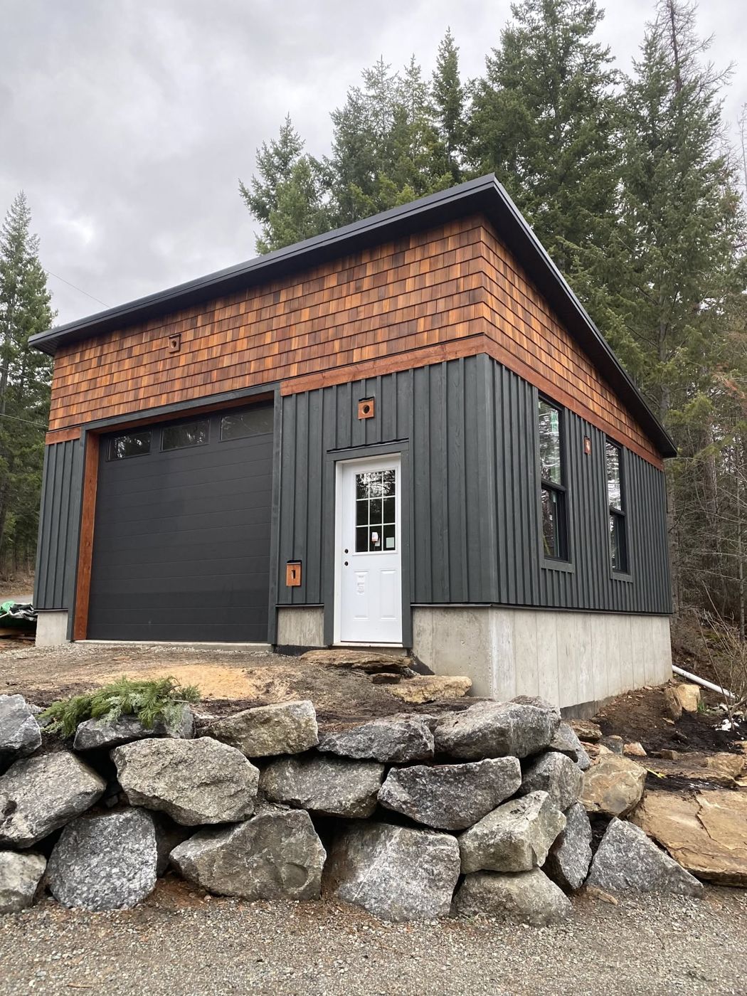 Shop with cedar shingle and dark board-and-batten siding on a concrete foundation, with a rock retaining wall